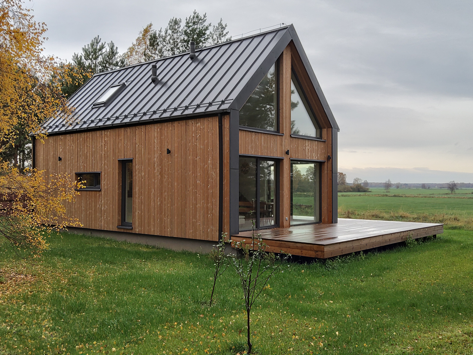 Drewniany dwupiętrowy dom szkieletowy, wykończony drewnem, z dużymi przeszkleniami i tarasem dookoła na zielonej działce, wśród jesiennych rdzawych drzew
