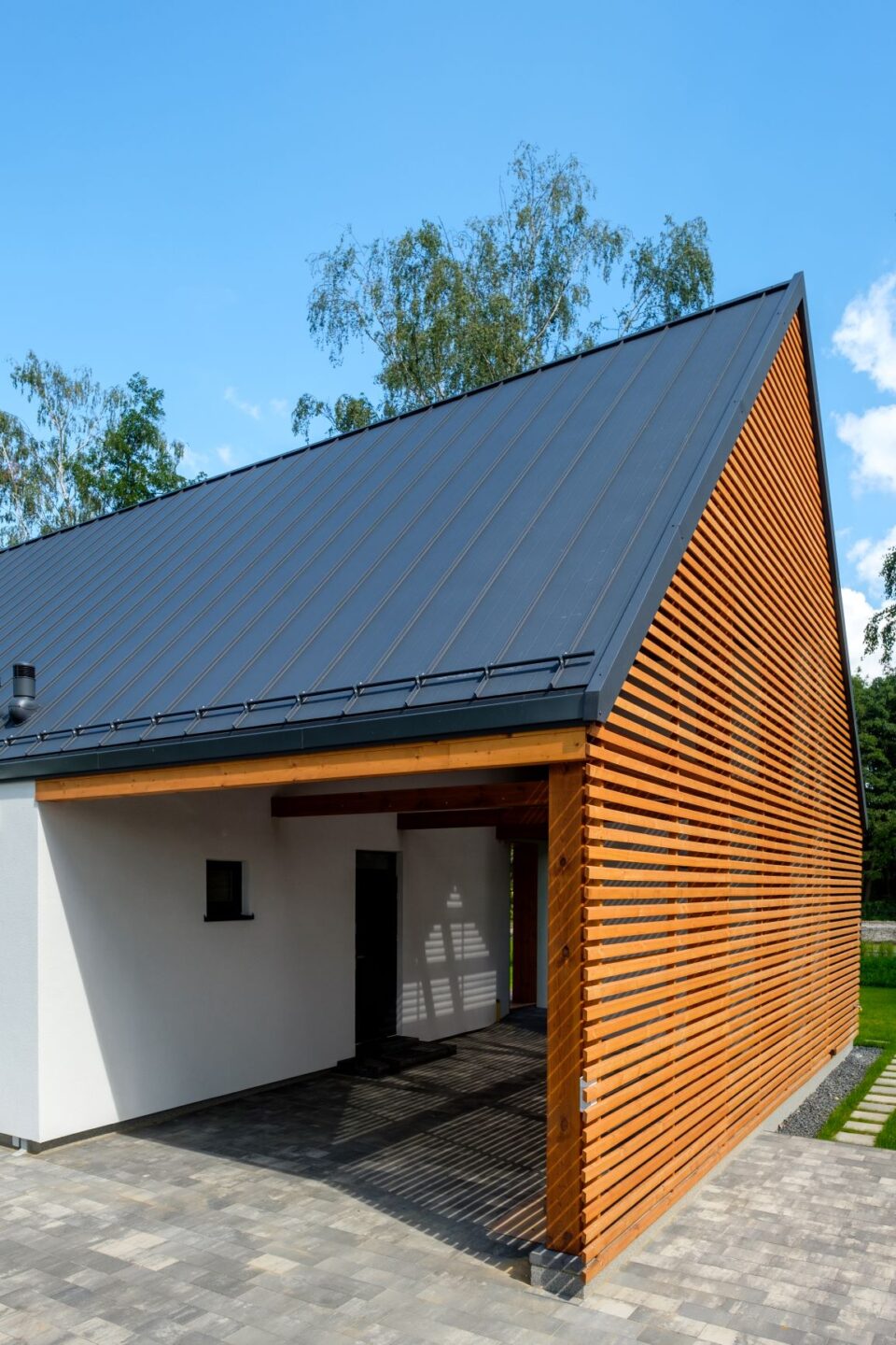 Dom szkieletowy Simple House, dobudowana drewniana wiata połączona z&nbsp;budynkiem otynkowanym na biało, pokryta dwuspadowym dachem z&nbsp;blachy antracytowej